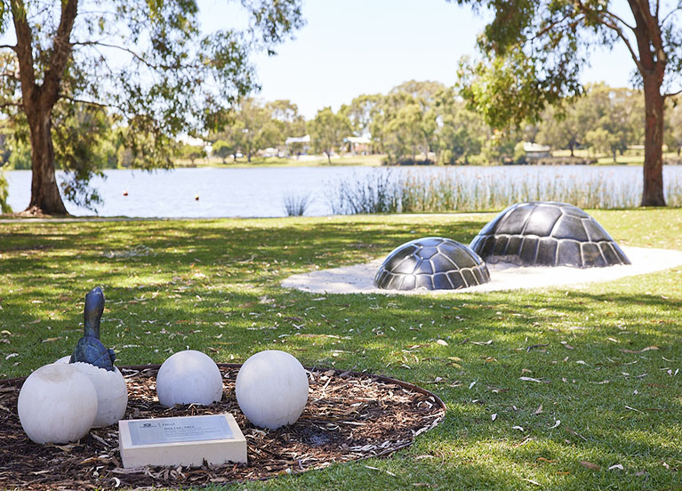Image of turtle family stone artwork including five eggs, with one hatching and two large granite shells. The view of Jackadder Lake in the background.