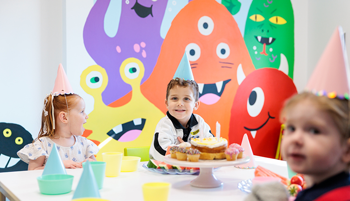 Children wearing party hats sit around a table with cupcakes in front of a colourful monster mural backdrop.