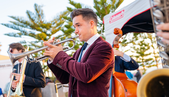 Musicians performing outdoors, with a trumpet player in the foreground and other jazz band members playing brass and string instruments in the background.