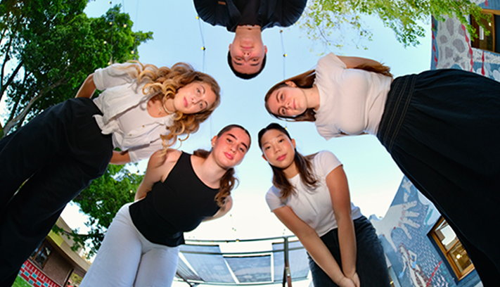 Five young performers viewed from below, standing in a circle outdoors and looking down toward the camera.