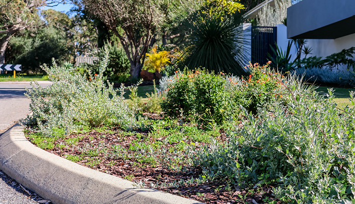 City worker standing beside a ride-on mower in a roadside reserve, with residential homes and trees in the background.