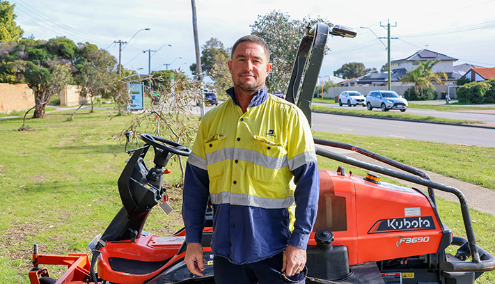 City worker standing beside a ride-on mower in a roadside reserve, with residential homes and trees in the background.
