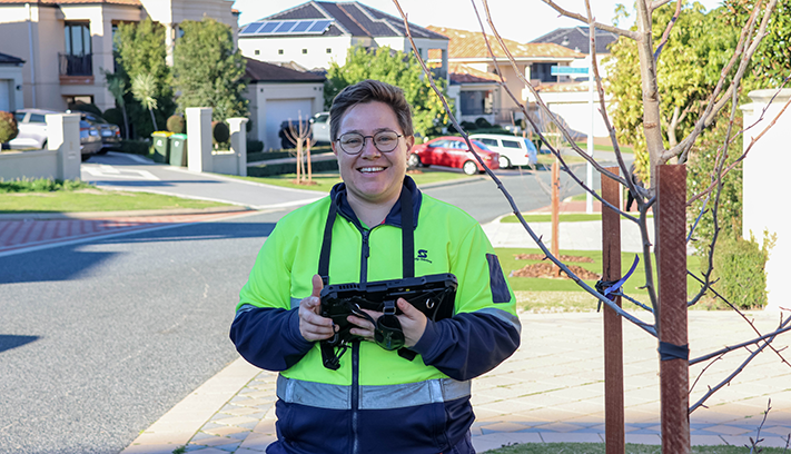 City worker holding a tablet beside a newly planted street tree in a residential neighbourhood.