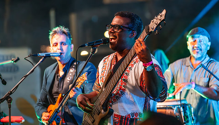 Musicians performing on stage at a live music event, with a singer playing bass guitar in the foreground and band members playing guitar and percussion in the background.