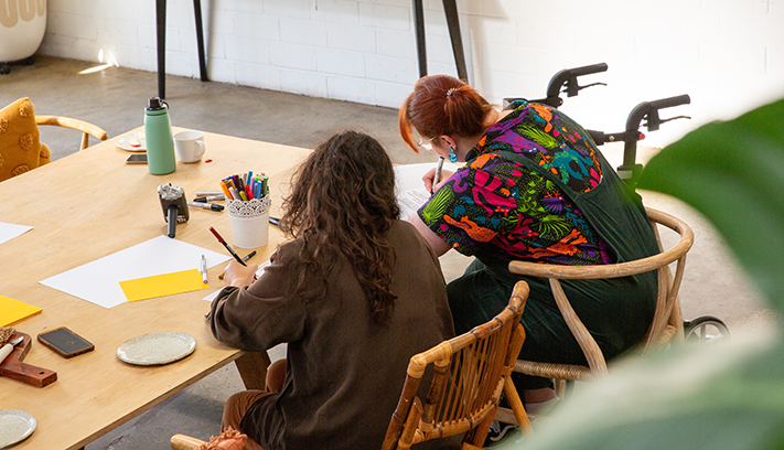 Two people seated at a table writing and drawing during a creative workshop, with art supplies and stationery spread across the workspace.