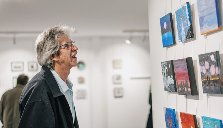Person viewing a display of small artworks on a gallery wall during an exhibition.