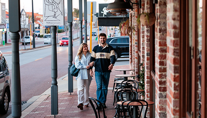 Two people walking along a shop-lined main street, with outdoor café seating and local businesses in the background.