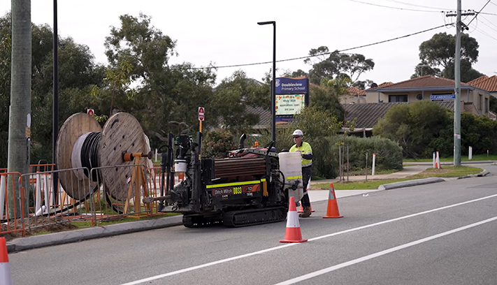 Roadworks crew operating underground drilling equipment beside a suburban street, with traffic cones and utility cable reels in place.