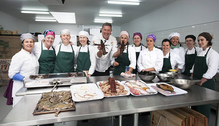 Group of culinary students and chefs standing behind a preparation table displaying seafood, including lobster, fish, crab and shellfish, in a commercial training kitchen.