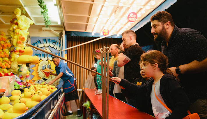 Child using a hook to catch rubber ducks at a carnival game stall, with families gathered around the attraction at night.