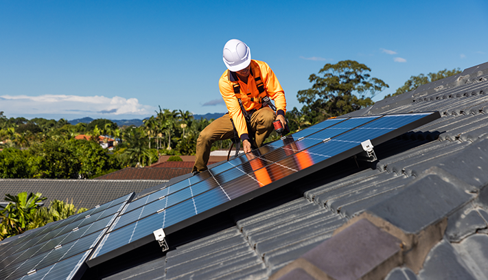Worker installing rooftop solar panels on a residential home, wearing safety equipment and using power tools.