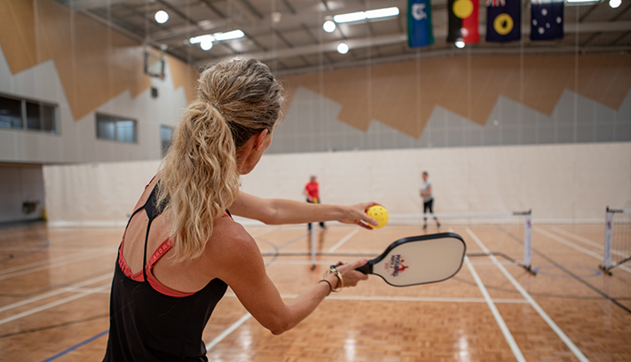 Person serving a pickleball during an indoor game on a sports court, with other players and multiple pickleball nets visible in the background.