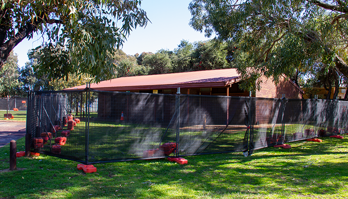 Temporary fencing surrounding a community building and adjacent grassy area during maintenance or construction works, with trees and parkland in the background.