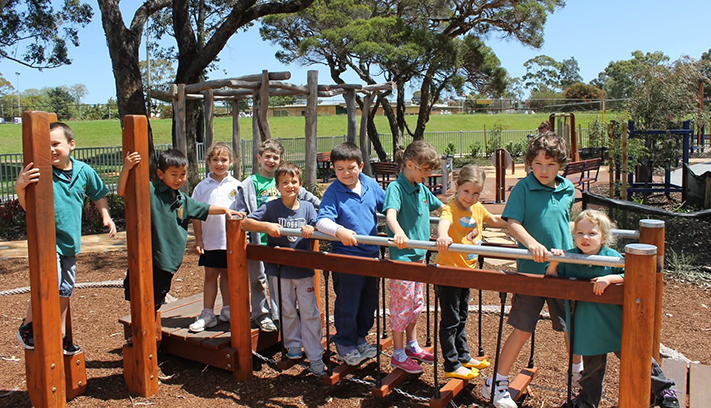 Children playing on a nature-based playground climbing structure, with trees, garden beds and open parkland in the background.