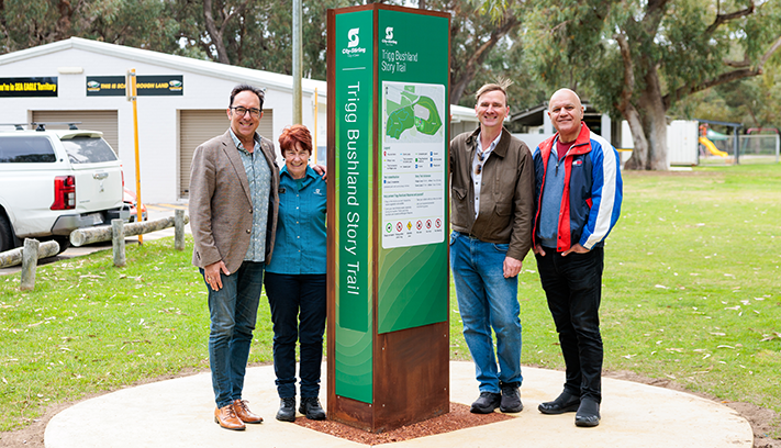 Four people standing beside a Trigg Bushland Story Trail information sign in a park, with trees and community facilities in the background.
