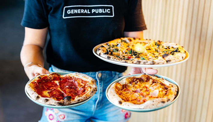 Three pizzas held by a waiter with a shirt that says GENERAL PUBLIC.