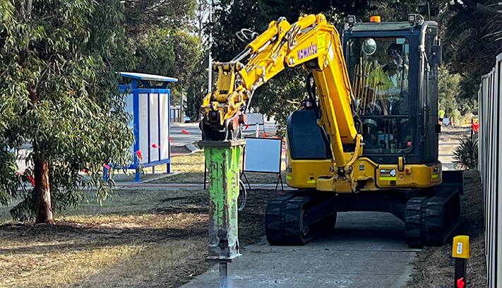 Excavator using a hydraulic breaker to remove a concrete footpath during construction works, with trees and a bus stop visible in the background.