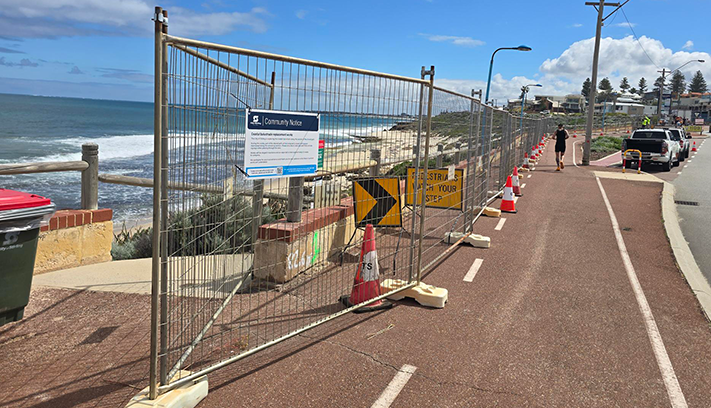 Temporary fencing and traffic cones installed along a coastal shared path during balustrade replacement works, with a community notice sign, ocean views and nearby roadway visible.