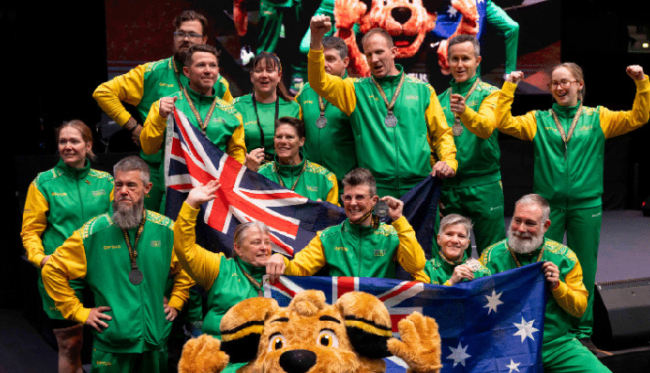 Group of athletes wearing green and gold tracksuits standing on a stage with medals and Australian flags, posing for a team photo at a sporting event.