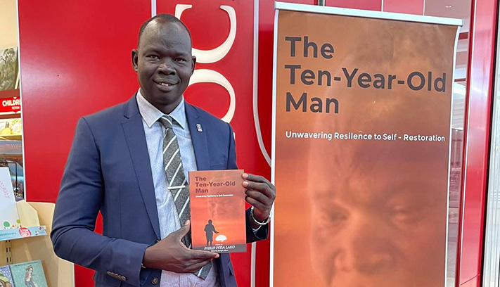 uthor holding a copy of The Ten-Year-Old Man beside a banner promoting the book at a library or community venue.