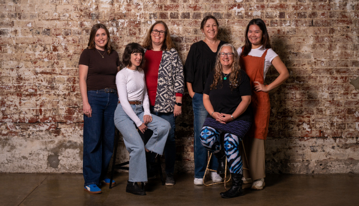 Six artists pose in a studio against a weathered brick wall, with one seated on a stool in front and others standing close behind in casual clothing.