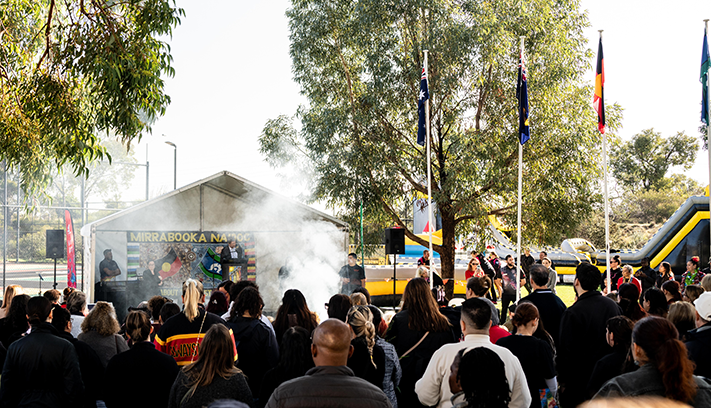 Crowd gathered at an outdoor NAIDOC Week community event, facing a stage with speakers, flags and a smoking ceremony.