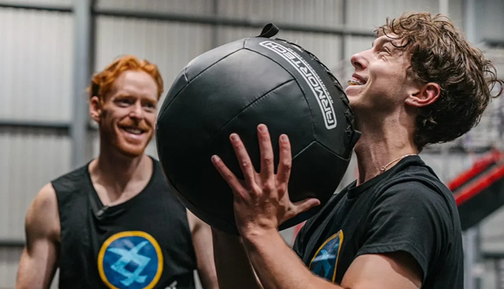 A person lifts up a medicine ball while a trainer watches on.