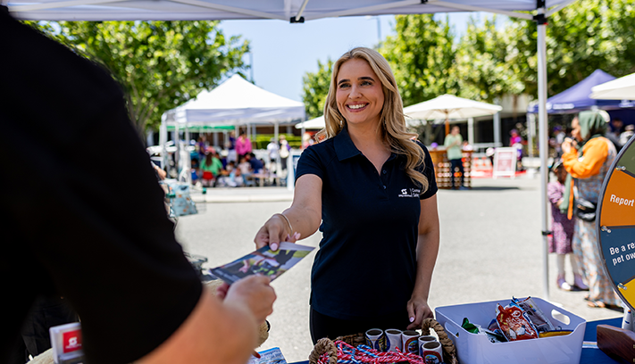 Person at a community market stall handing an information flyer to a visitor under a marquee.
