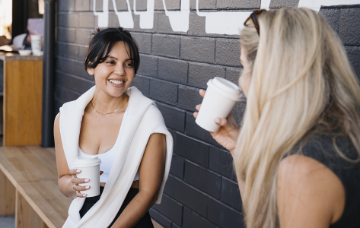 Two women share a cup of coffee on a bench.
