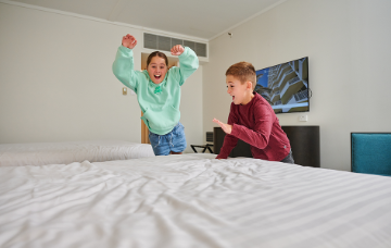 Two children excitedly jump onto a bed in a hotel room.
