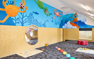 Children’s activity room with tables, soft seating and a colourful monster mural painted across one wall.