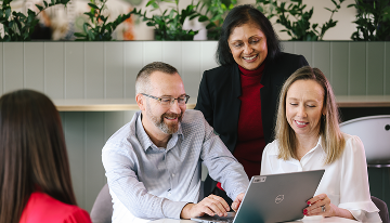 Three people gathered around a laptop in an office setting, reviewing information together at a table.