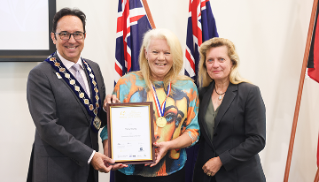 Three people standing together during an award presentation, holding a framed certificate and medal in front of Australian and Aboriginal flags.