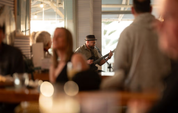 Musician performing with a guitar and microphone in a café or bar, with patrons visible in the foreground.