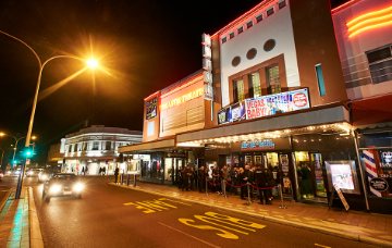 The Astor Theatre at night, with people queuing outside the illuminated cinema entrance along a busy street.