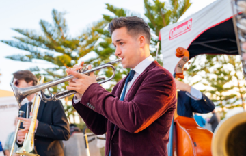 Musicians performing at an outdoor jazz event, with a trumpet player in the foreground and other band members playing brass and string instruments in the background.