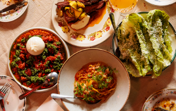 Overhead view of shared restaurant dishes, including pasta, salad, burrata and roasted meat, arranged on a dining table.