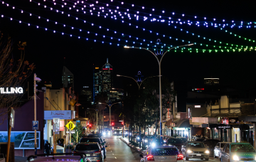 Night-time view of a busy main street illuminated by colourful overhead light displays, with shops, traffic and the city skyline in the background.