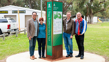 Four people standing beside a Trigg Bushland Story Trail information sign in a park, with trees and community facilities in the background.