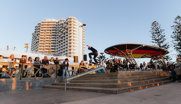 Skateboarder performing a trick down a handrail beside a staircase at Scarborough Beach, with spectators gathered around the public plaza and beachfront buildings in the background.