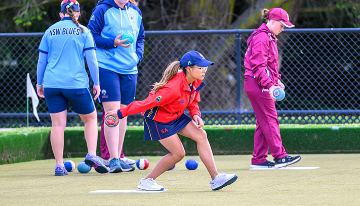 Young lawn bowls players competing on a bowling green, with a player delivering a bowl while other participants and bowls are visible in the background.