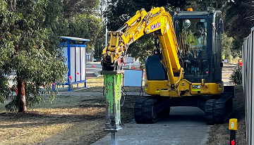 Excavator using a hydraulic breaker to remove a concrete footpath during construction works, with trees and a bus stop visible in the background.