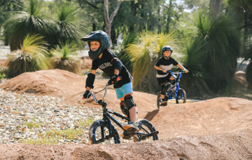 Two children wearing helmets ride bicycles along a pump track surrounded by native vegetation.