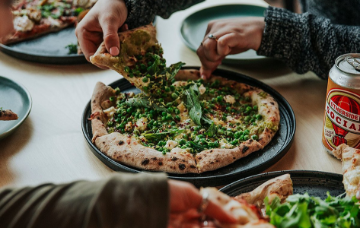 Hands reaching for slices of pizza topped with greens and cheese, shared around a restaurant table with plates and drinks.