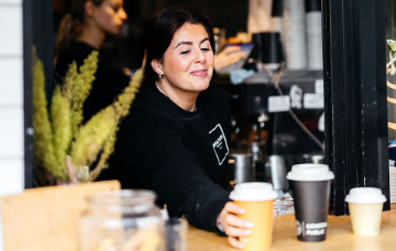 Coffee shop staff member handing a takeaway coffee to a customer at a café service window, with coffee cups and equipment visible in the background.