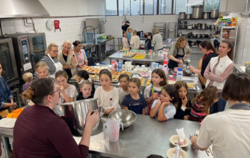 Children and adults gathered around a workbench in a commercial kitchen, taking part in a hands-on cooking workshop.