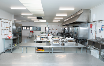 Commercial training kitchen with stainless steel workstations, industrial ovens and food preparation areas, with culinary students working in the background.