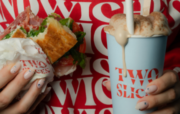 Person holding a gourmet sandwich and a takeaway drink topped with toasted marshmallow foam, with Two Slices branding visible in the background.